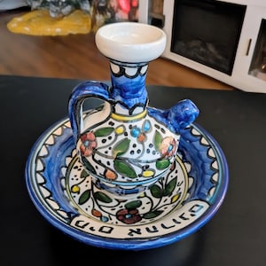 May include: A ceramic hand-washing set consisting of a small pitcher with a handle and spout and a matching round basin. The set features a white base with hand-painted blue, green, and red floral patterns and Hebrew text along the basin rim.