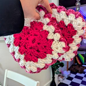 May include: Two crocheted squares in pink and white, held in a hand. The squares are made with a granny stitch pattern.