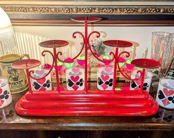 May include: A red metal candle holder with six circular platforms for candles. The holder has a decorative design with heart-shaped accents and sits on a wooden surface. The candle holders are accompanied by decorative glasses.