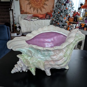 May include: A large ceramic conch shell planter featuring a glossy, iridescent exterior with subtle green and white textured ridges. The interior of the shell is finished in a smooth, vibrant lavender glaze with a pearlescent sheen.