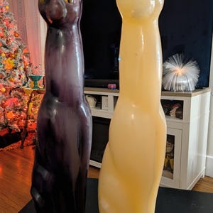 May include: Two tall, slender cat-shaped candles standing upright. The candle on the left is a deep, dark purple with amber-colored eyes. The candle on the right is a pale, creamy off-white color with bright green eyes. Both have pointed ears.