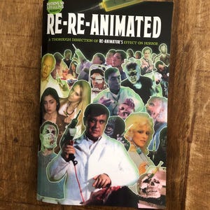 May include: A book titled "RE-RE-ANIMATED" with a collage of horror movie imagery. The cover features the text "A THOROUGH DISSECTION OF RE-ANIMATOR'S EFFECT ON HORROR". The book is on a wooden surface.