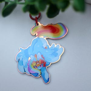 May include: A decorative acrylic keyring featuring a light blue winged pony character with a rainbow-coloured mane and tail. The character wears round goggles and is depicted in a dynamic pose. The charm is attached to a small red metal ring.
