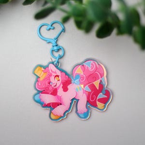 May include: A light blue heart-shaped lobster clasp keyring attached to an acrylic charm featuring a pink pony character with curly hair, a yellow top hat, and a large, flowing pink and blue tail with yellow triangular accents on a clear base.