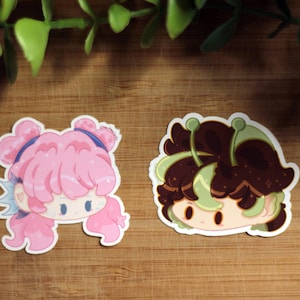 May include: Two cartoon stickers on a wooden surface. One sticker features a pink-haired character with blue accents. The other sticker depicts a character with brown hair and green accents. Both stickers have a white border.