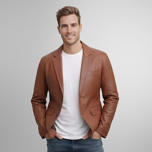 Puede incluir: Una blazer de cuero marrón con cierre clásico de dos botones, un bolsillo en el pecho y dos bolsillos delanteros. La blazer se lleva sobre una camiseta blanca. El modelo sonríe, con las manos en los bolsillos.