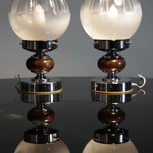 Może przedstawiać: Para lamp stołowych w stylu Art Deco. Każda lampa ma klosz w kształcie kuli z matowego szkła z przezroczystym, żebrowanym wierzchem. Lampy mają chromowaną podstawę, ciemny drewniany akcent i mały uchwyt. Lampy stoją na odblaskowej powierzchni.