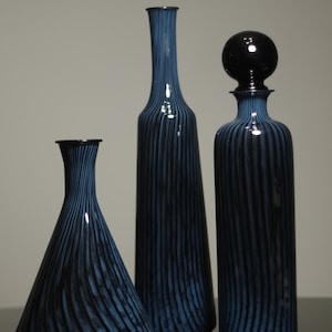 Peut inclure: Trois bouteilles en verre bleu avec des rayures noires verticales. Les bouteilles varient en forme et en hauteur, l'une d'elles étant munie d'un bouchon rond. Les bouteilles sont disposées sur une surface réfléchissante.