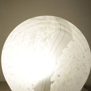 Może przedstawiać: Biała szklana lampa stołowa o wirującym, fakturowanym wzorze. Lampa jest oświetlona, dając ciepłe światło.