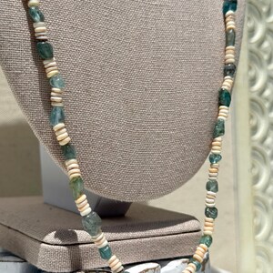 May include: A long necklace featuring a mix of seafoam green and off-white beads. The necklace is composed of irregular, square-shaped green beads interspersed with round, off-white shell beads. The necklace is displayed on a neutral-toned mannequin.