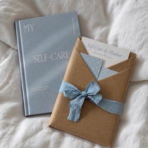 Peut inclure: Un journal à couverture rigide bleu clair intitulé MY SELF-CARE A SANCTUARY FOR MY SOUL, MIND & BODY à côté d'un paquet enveloppé de papier kraft brun noué d'un ruban bleu clair, contenant une carte Self-Care & Mindset Journal Prompts.