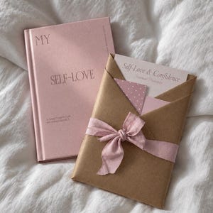 Op de afbeelding: Een roze hardcover dagboek met de titel MY SELF-LOVE A SANCTUARY FOR MY INNER WORLD naast een bruine papieren envelop met een roze lint, met daarin een kaart met de tekst Self-Love & Confidence Journal Prompts en een klein roze papier met stippen.