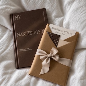 Może przedstawiać: Ciemnobrązowa książka w twardej oprawie z napisem MY MANIFESTATION A SANCTUARY FOR INTENTION & ALIGNMENT obok owiniętego w brązowy papier pakunku przewiązanego beżową wstążką, zawierającego kartkę z napisem Manifestation & Success Journal Prompts.