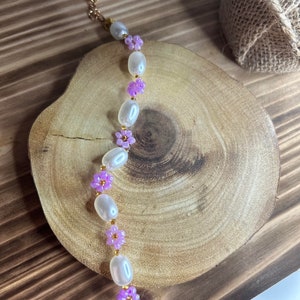 May include: A delicate bracelet featuring white oval beads and small purple flower accents. The bracelet is made with gold-toned metal.