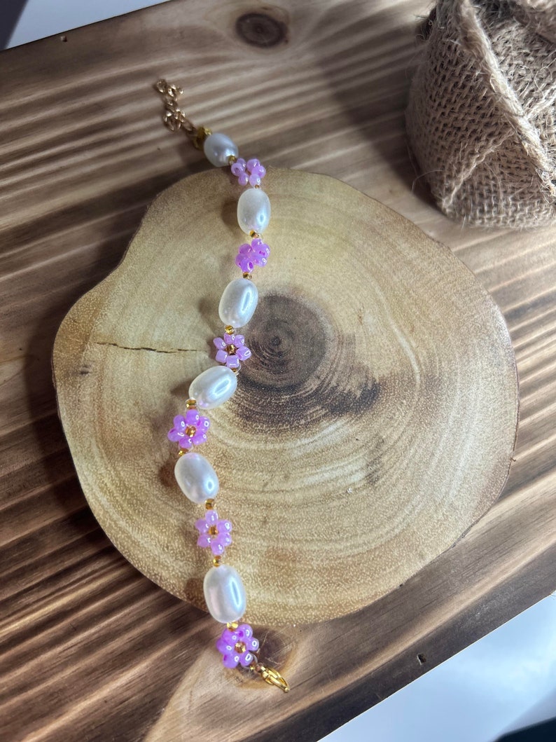 May include: A delicate bracelet featuring white oval beads and small purple flower accents. The bracelet is made with gold-toned metal.