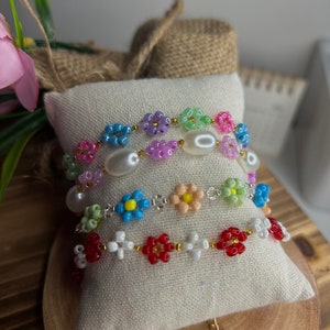 May include: Three beaded bracelets with flower-shaped beads. The top bracelet is multi-colored with pink, green, blue, and purple beads. The middle bracelet is white with blue, green, and peach flower beads. The bottom bracelet is white with red flower beads.