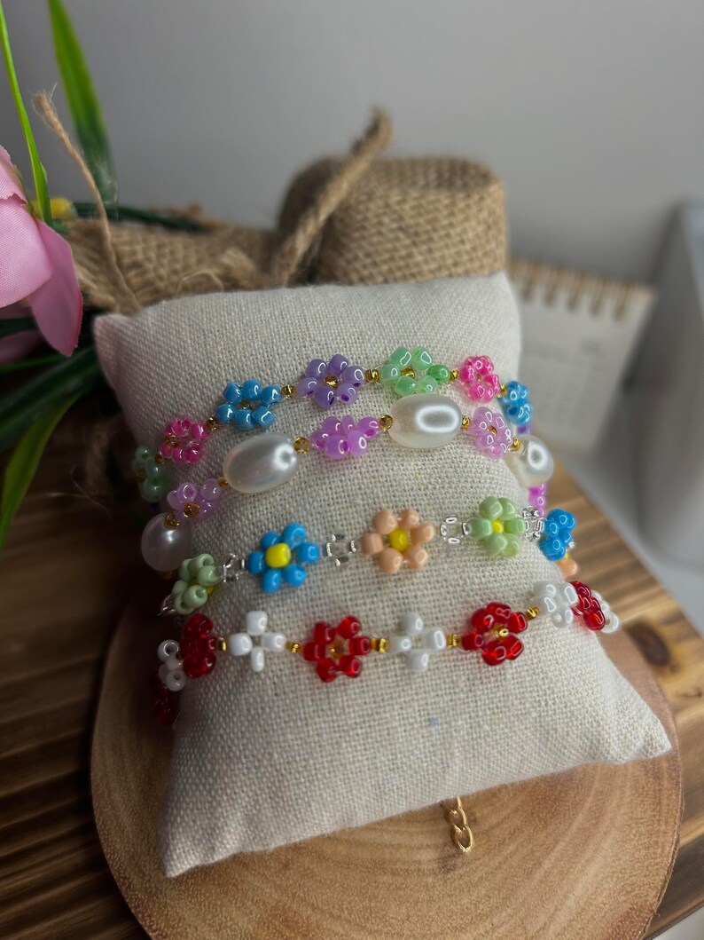 May include: Three beaded bracelets with flower-shaped beads. The top bracelet is multi-colored with pink, green, blue, and purple beads. The middle bracelet is white with blue, green, and peach flower beads. The bottom bracelet is white with red flower beads.