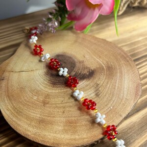 May include: A red and white beaded bracelet with flower-shaped beads. The bracelet is on a wooden surface.
