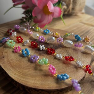 May include: Three colorful beaded necklaces with flower-shaped beads. The necklaces are made with white, pink, blue, green, red, and purple beads. The necklaces are on a wooden surface.