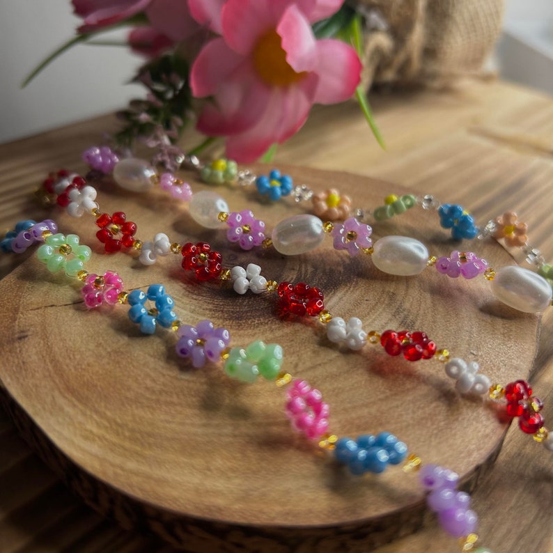 May include: Three colorful beaded necklaces with flower-shaped beads. The necklaces are made with white, pink, blue, green, red, and purple beads. The necklaces are on a wooden surface.