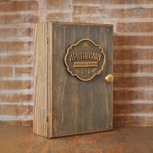 Corner view of rustic wooden apothecary-style box with bronze and gold accents with 'Apothecary Potions & Elixirs' label on a wooden background