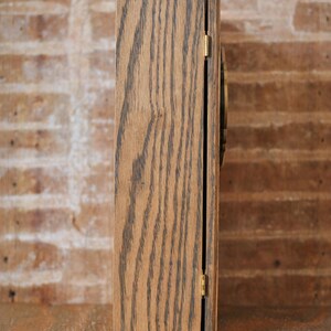 May include: A rectangular wooden box viewed from the side, featuring a hinged door. The box is crafted from light brown wood with prominent dark grain. A small gold hinge secures the door. The backdrop is a brick wall.