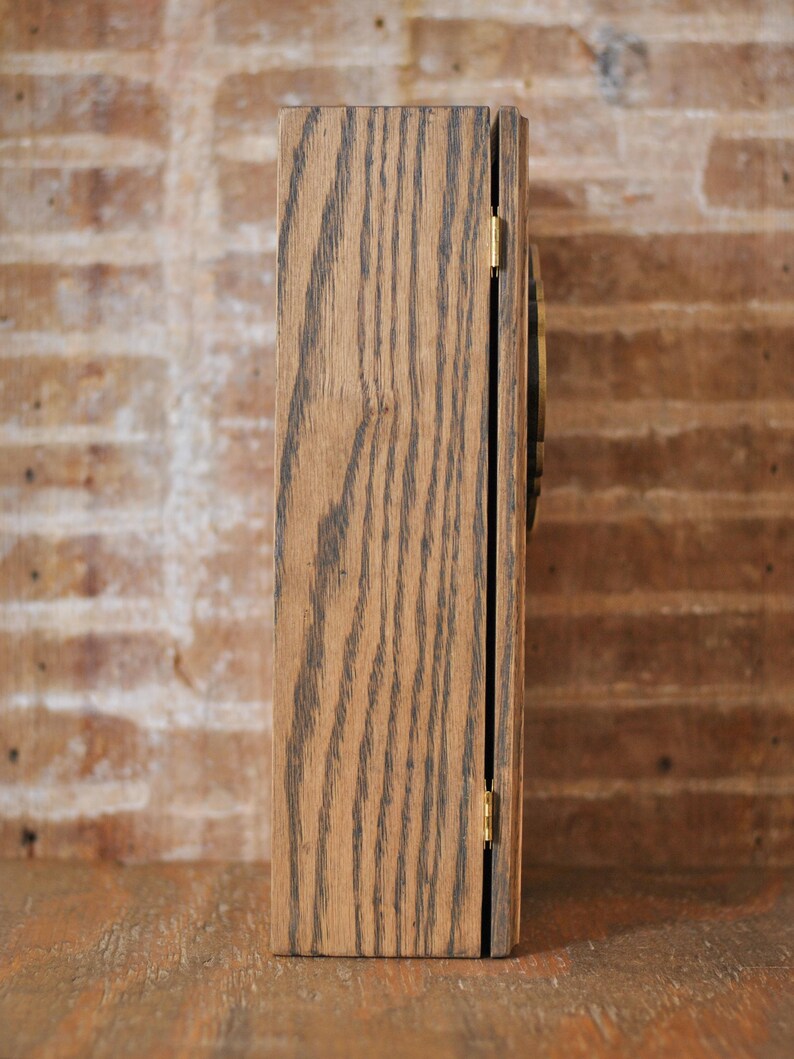 May include: A rectangular wooden box viewed from the side, featuring a hinged door. The box is crafted from light brown wood with prominent dark grain. A small gold hinge secures the door. The backdrop is a brick wall.