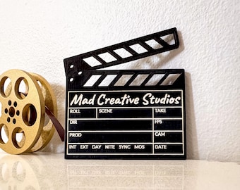 May include: A black wooden clapperboard with the text "Mad Creative Studios" and film reel. The clapperboard has white text and is open. The film reel is gold-colored.