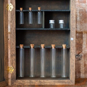 rustic wooden apothecary-style box with bronze and gold accents, glass vials and jars in various sizes with 'Apothecary Potions Elixirs' label on a wooden tabletop and brick background