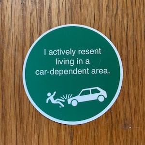 Könnte beinhalten: Ein grüner runder Aufkleber mit weißem Text: „Ich lehne es aktiv ab, in einem autoabhängigen Gebiet zu leben.“ Der Aufkleber zeigt auch eine weiße Grafik einer Person, die von einem Auto angefahren wird.