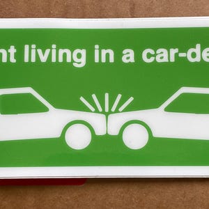 Könnte beinhalten: Ein grüner und weißer Aufkleber mit dem Text "Ich ärgere mich aktiv darüber, in einem autoabhängigen Gebiet zu leben." Zwei weiße Autos sind dargestellt, die miteinander kollidieren.