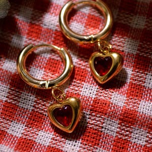 May include: A pair of gold hoop earrings with dangling red heart charms. The earrings are on a red and white checkered background.