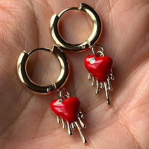 May include: A pair of gold hoop earrings with red enamel heart charms. The hearts have a small diamond accent and dripping red enamel detail.