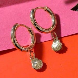May include: Gold hoop earrings with a small seashell charm hanging from each hoop. The earrings are on a pink and orange background.