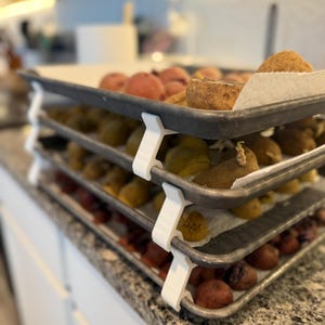 May include: A three-tiered tray system with white supports, holding various fruits and baked goods. The top tray features peaches, the middle tray has yellow fruits, and the bottom tray displays red fruits. The trays are on a granite countertop.