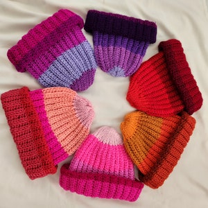Puede incluir: Siete gorros de ganchillo dispuestos en círculo, cada uno con un degradado de color diferente. Los gorros están hechos con un hilo grueso y tienen un estilo sencillo y holgado. Los colores son morado, rojo, naranja, rosa, melocotón, lavanda y rojo.