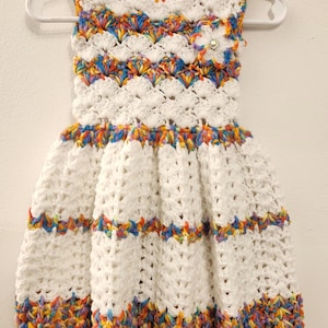 May include: A white crochet dress with a sleeveless design, featuring colorful rainbow accents at the neckline, waist, and hem. A decorative flower with a rhinestone center is attached near the shoulder. The dress is displayed on a white hanger.