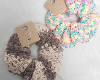 May include: Two handmade crochet scrunchies. One is a mix of pastel colors: pink, yellow, and blue. The other is a mix of brown and beige. Each scrunchie has a small tag that says "Handmade with love".