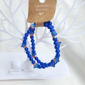 May include: Two blue beaded bracelets with star and flag accents, displayed on a white decorative tree. The bracelets are strung with small, round beads and feature star-shaped charms with red, white, and blue flag designs. A brown tag reads "Handmade with love."