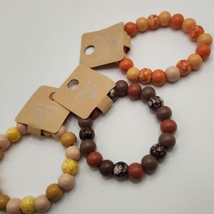 May include: Three stretch bracelets featuring round beads in shades of orange, brown, and yellow. Each bracelet is attached to a rectangular tan cardboard tag with the text Handmade with love printed in white and a small heart icon visible.