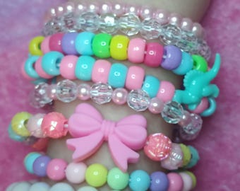 May include: A collection of colorful beaded bracelets stacked on a wrist. The bracelets feature a variety of beads in pastel colors, including pink, blue, yellow, and purple. Some bracelets have clear beads, a pink bow, and a turquoise dinosaur charm.