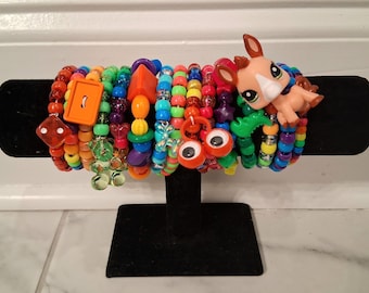 May include: A collection of colorful beaded bracelets displayed on a black stand. The bracelets feature a variety of beads in rainbow colors, with charms including an orange button, dice, and a cartoon animal figure. The bracelets are a fun and playful accessory.