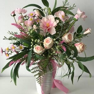 Puede incluir: Un arreglo floral en un jarrón blanco. El ramo presenta rosas rosas, amapolas y otras flores con follaje verde. Una cinta rosa está atada alrededor del jarrón. El arreglo está sobre un fondo blanco.