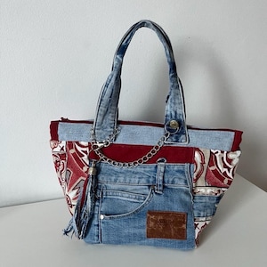 Może przedstawiać: Torba typu tote z dżinsu z patchworkowym wzorem. Torba ma czerwoną podszewkę i boczne panele we wzory. Srebrny łańcuszek i frędzel dodają szczegółów. Torba ma dżinsową kieszeń i brązową skórzaną naszywkę.