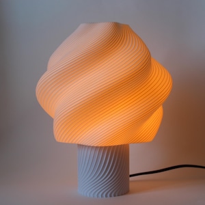 Pode incluir: Uma lâmpada de mesa branca e moderna com um design único em espiral. A lâmpada tem uma base cilíndrica e um topo bulboso, ambos com uma textura canelada. O interior brilha com uma luz laranja quente, e um cabo de alimentação preto estende-se da base.