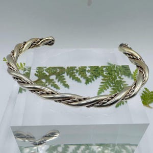 May include: A silver-toned cuff bracelet featuring a twisted design. The bracelet is constructed from a thicker, smooth twisted metal strand intertwined with a thinner, rope-textured metal strand, creating a continuous spiral pattern.