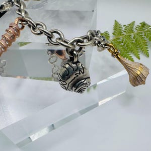 May include: A silver-toned metal charm bracelet featuring a thick chain link design. The bracelet includes three distinct charms: a rose gold-toned coiled cylinder with embedded crystals, a round silver shell charm, and a gold-toned teardrop pendant.