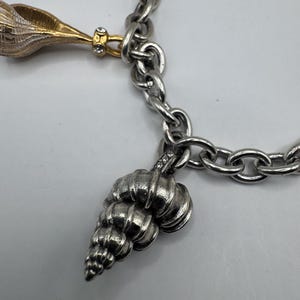 May include: A silver-toned link charm bracelet featuring multiple hanging charms. One charm is a gold-toned seashell with small clear stones, and another is a textured silver-toned seashell charm with small clear stones at the top attachment point.