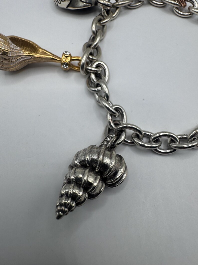 May include: A silver-toned link charm bracelet featuring multiple hanging charms. One charm is a gold-toned seashell with small clear stones, and another is a textured silver-toned seashell charm with small clear stones at the top attachment point.