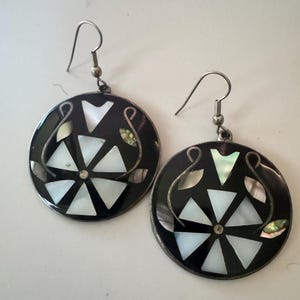 May include: A pair of round, black earrings with a silver hook. Each earring features a central design of white triangles and iridescent accents, surrounded by a silver border. The earrings have a unique, geometric pattern.
